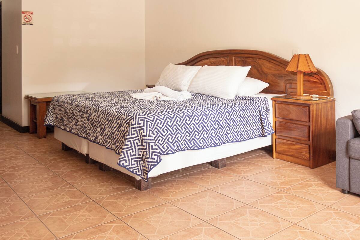 A bedroom with a bed, a wooden nightstand, a lamp, a ceiling fan, and a tiled floor.