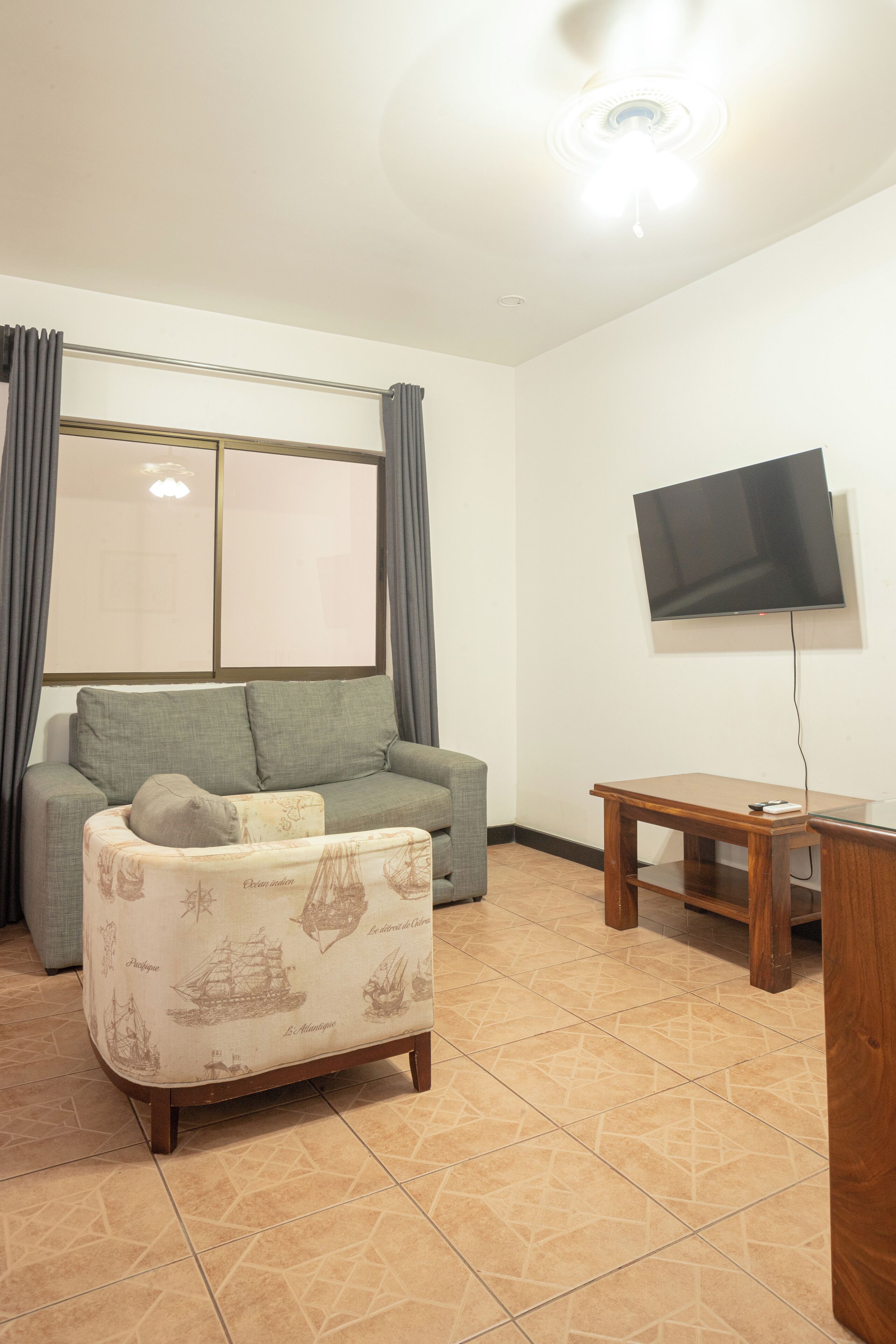 A living room with a sofa, a small table, a flat-screen TV mounted on the wall, and a window with curtains.