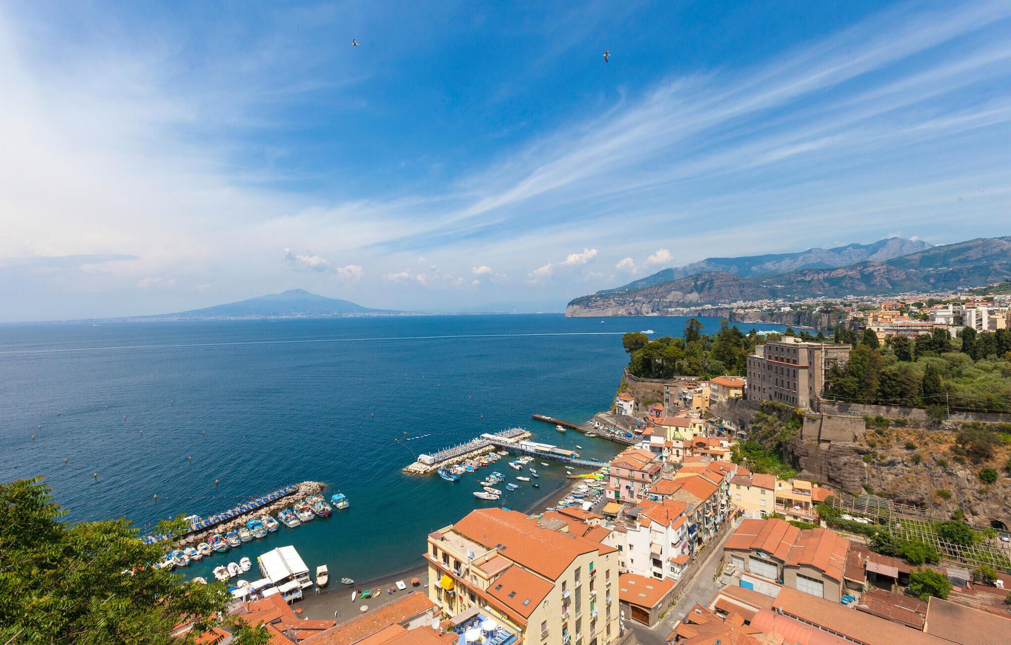 Una cittadina costiera con un porto turistico, edifici dai tetti rossi e montagne sullo sfondo.