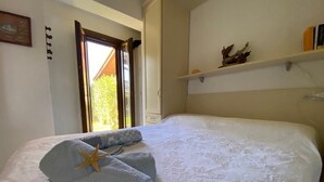 Una cama bien hecha con un adorno de estrella de mar, una ventana con vistas a la vegetación y un estante con objetos decorativos.