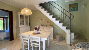 Un comedor con una mesa y sillas blancas, una lámpara colgante, una escalera con barandilla de metal y un armario con puertas de vidrio.
