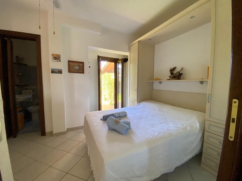 Un dormitori amb un llit, una porta, una estant amb objectes decoratius, i una vista a través d'una porta oberta.