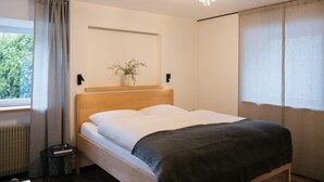A modern hotel room with a wooden bed, a bedside table with a book, a small plant, and a view of greenery outside.