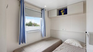 A single bed with a headboard, a window with curtains, a shelf with a vase, and a wall-mounted light fixture. - Mobile Home "Villa Granja Sol 2" with Shared Garden & Wi-Fi (São Felix da Marinha)