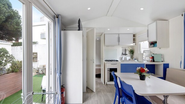 A modern kitchen with white cabinets, a dining area with blue chairs, and a view of a green garden through a sliding door. - Mobile Home "Villa Granja Sol 2" with Shared Garden & Wi-Fi (São Felix da Marinha)
