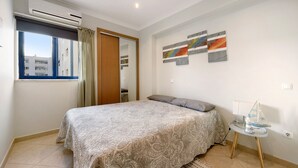 A bedroom with a bed, a window with curtains, a wall-mounted air conditioner, and a small table with a decorative item. - Apartment "Clube Praia da Rocha 3" with Balcony & Wi-Fi (Portimao)