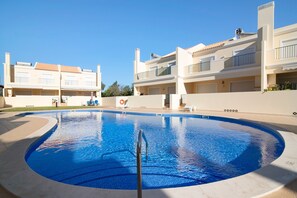 Une piscine à l’eau d’un bleu cristallin, entourée d’une terrasse carrelée et jouxtant un bâtiment doté de balcons et de fenêtres.