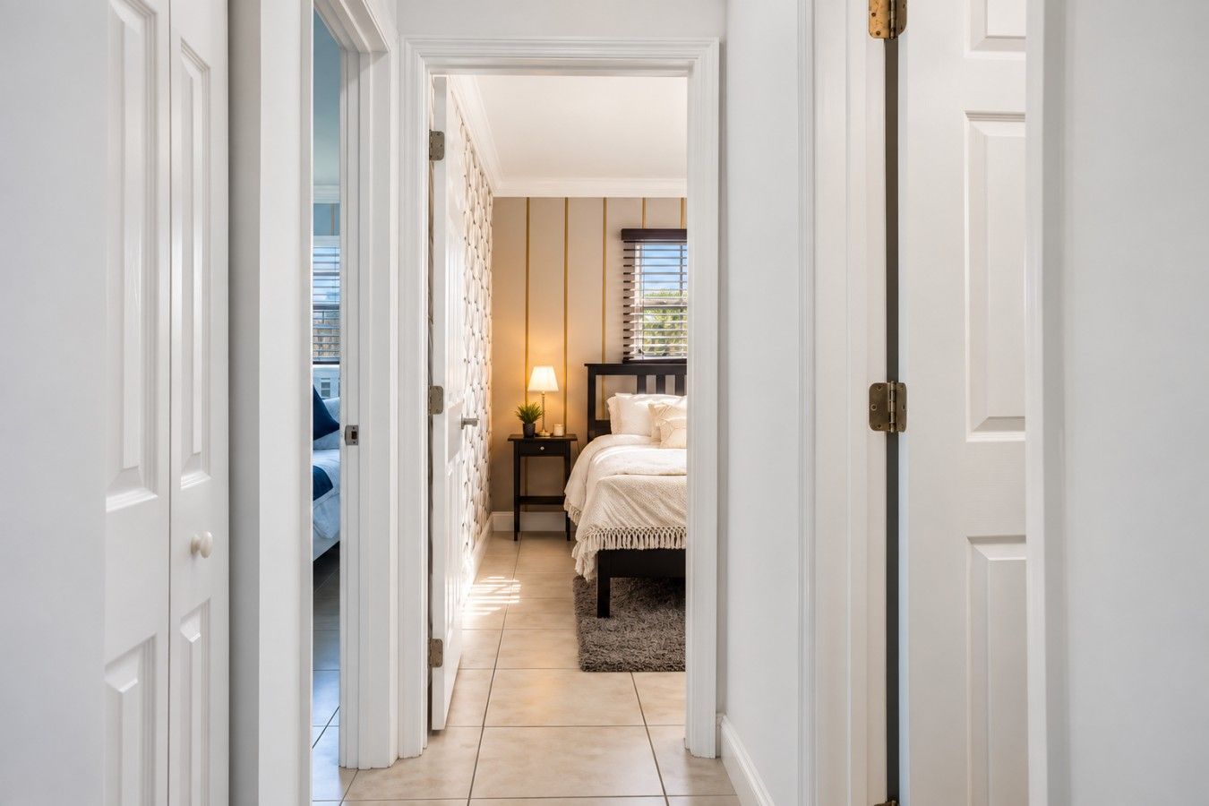 A hallway leading to a bedroom with a bed, bedside table, lamp, and window with blinds.