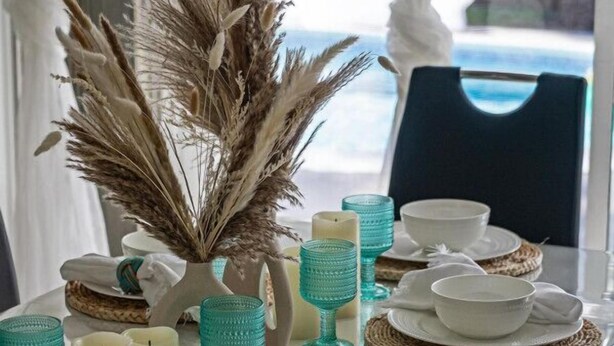 Une table à manger dressée avec de l’argenterie blanche, de la verrerie turquoise et des bougies, agrémentée d’une composition décorative de plantes séchées au centre.