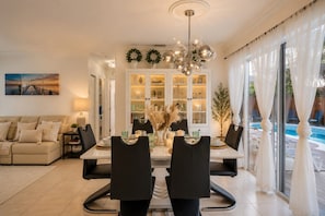 A dining area with a table set for two, a chandelier, and a view of a swimming pool through large windows. - Family Fun Villa | Heated Pool | Arcade & Ping Pong | BBQ | Near Hollywood Beach (Hollywood)