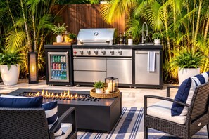 An outdoor kitchen with a grill, a refrigerator, and a built-in sink. There are wicker chairs and a fire pit.