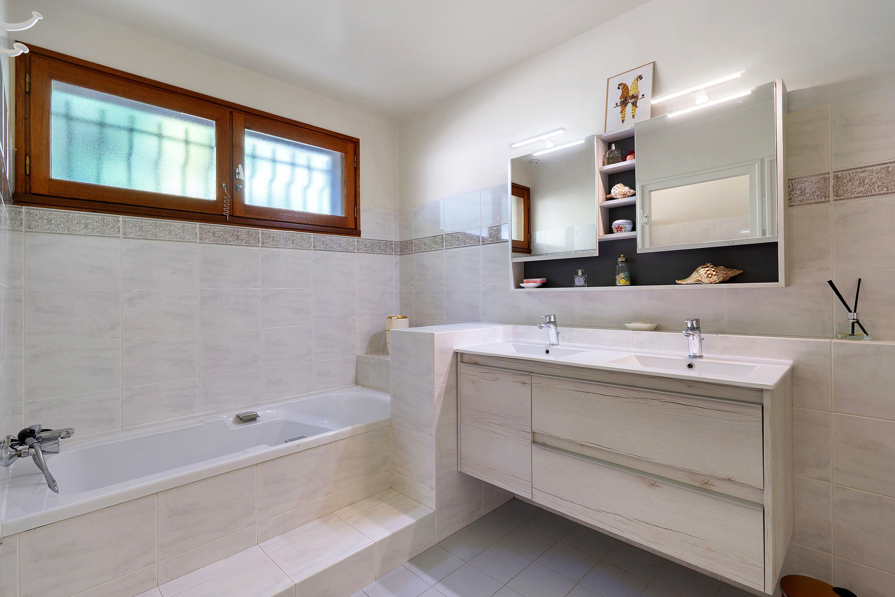 Uma casa de banho moderna com banheira, duas pias, toucador com gavetas, janela e espelho com prateleiras acima.