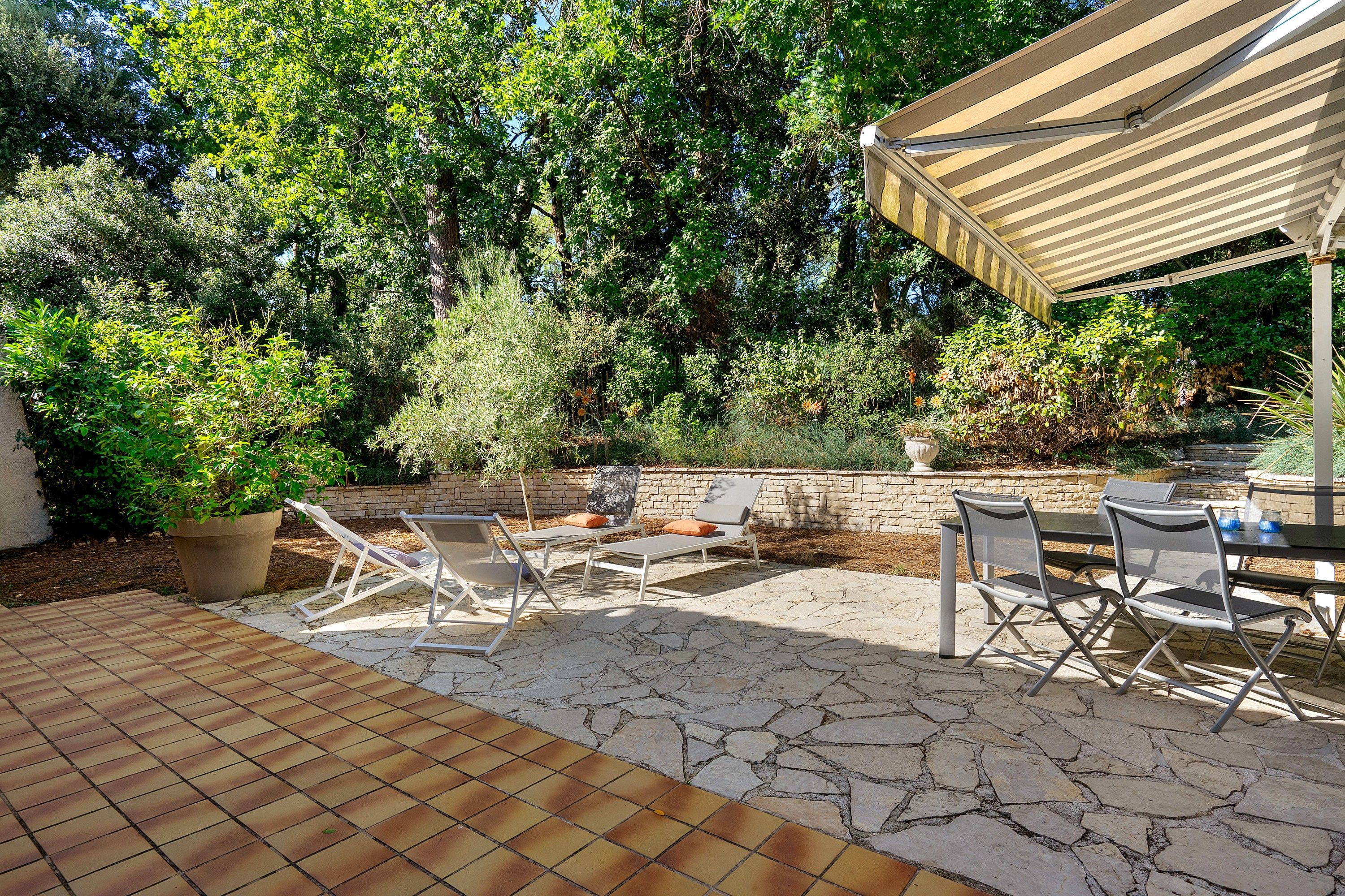 An outdoor patio with stone flooring, a covered seating area, and a variety of plants and trees.