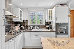 A modern kitchen with white cabinets, a stainless steel range hood, a double oven, and a window with heart-patterned curtains. - Chalet "Ma Bonne Etoile" with Mountain View, Private Terrace and Wi-Fi (Méolans-revel)