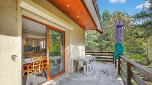 Een overdekt terras met een tafel en stoelen, een houten leuning en uitzicht op een bebost gebied.