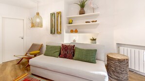 Un canapé blanc agrémenté de coussins verts et à motifs, une chaise en bois, un tabouret en osier et une étagère murale décorée de divers objets.