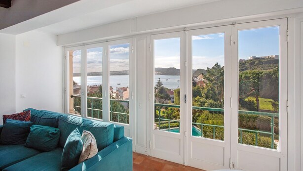 A room with a teal sofa, large windows, and a view of a body of water and greenery. - Vacation Home "São Martinho All The Best" with Sea View, Shared Pool & Wi-Fi (São Martinho do Porto)