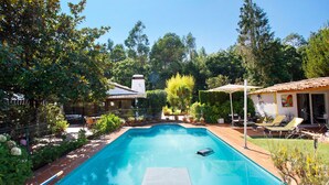 Una piscina con trampolín, rodeada de follaje y una casa con techo de tejas.