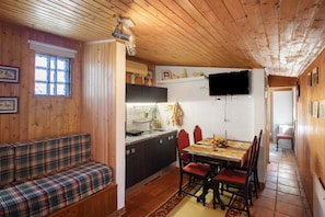 Interior acolhedor de uma cabana com teto de madeira, área de jantar com mesa e cadeiras, cozinha com pia e fogão, e janela com persianas.