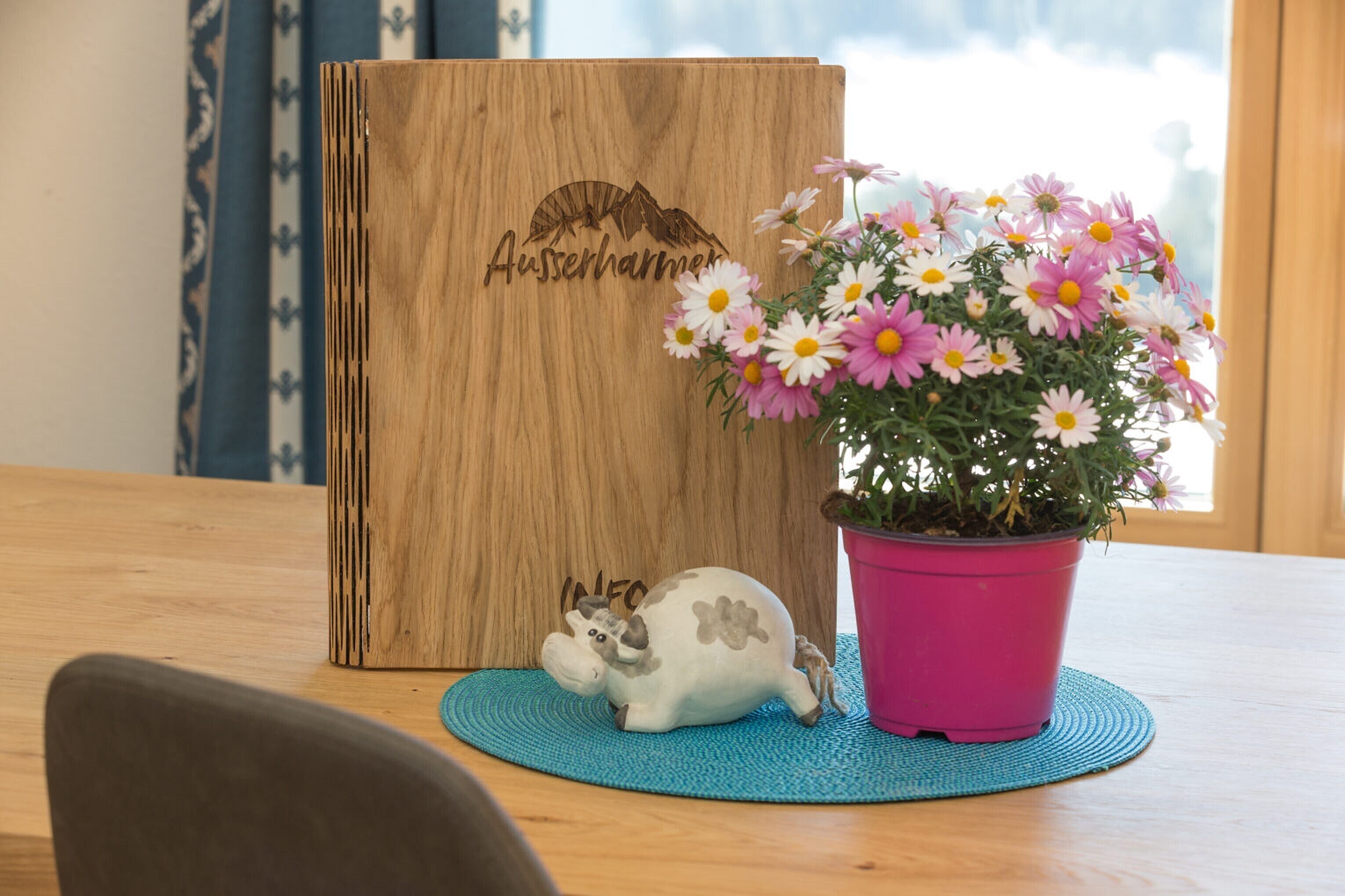 Une planche en bois portant l’inscription « Auserharmer », un cochon en céramique, une plante en pot avec des fleurs et une nappe bleue sur une table en bois.