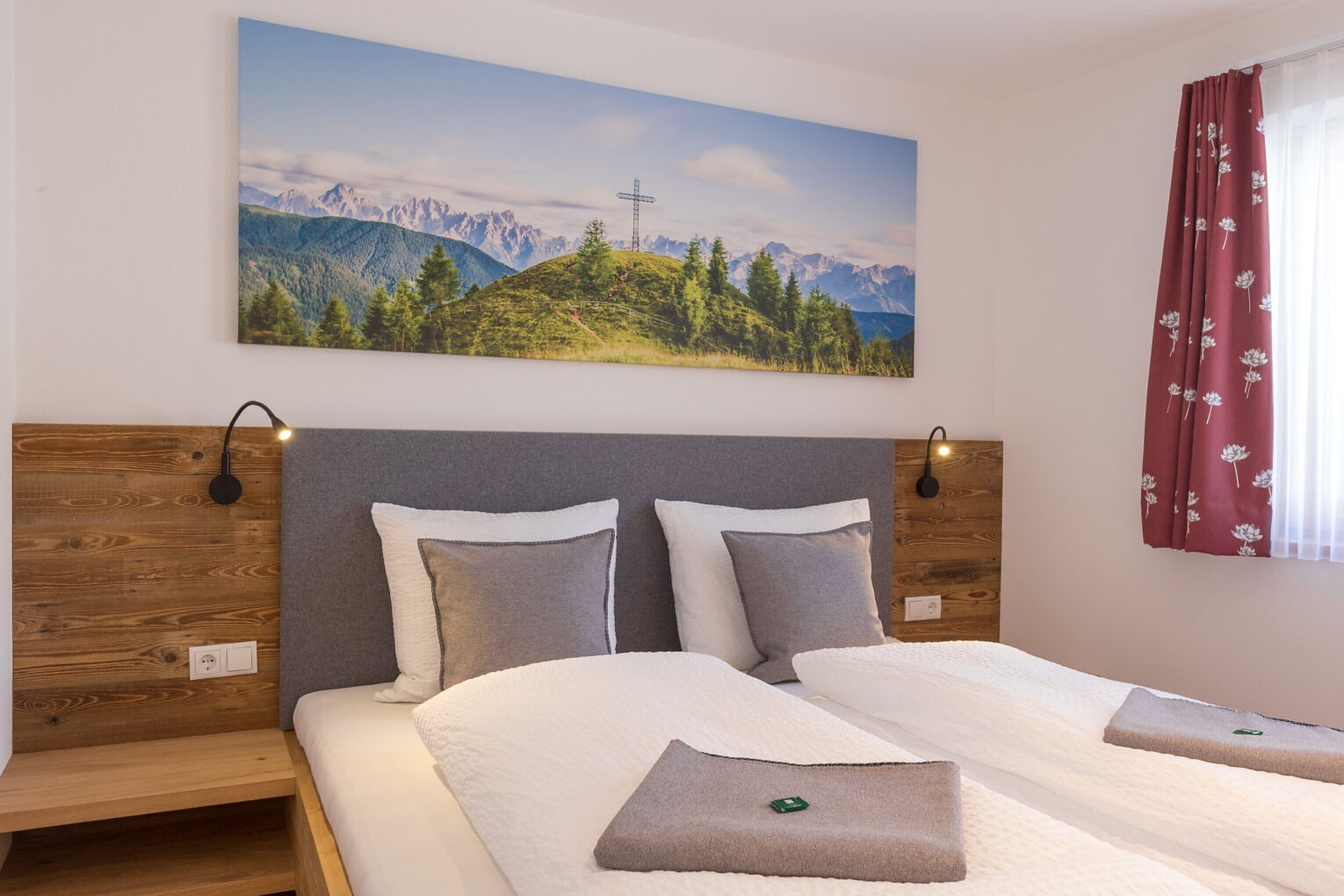 Habitación de hotel con cama, mesita de noche, lámpara de pared y un cuadro de un paisaje montañoso.