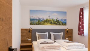 Un letto rifatto con cuscini e un asciugamano piegato, una testiera in legno, una lampada a parete e un dipinto che raffigura un paesaggio montano sopra il letto.