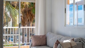 A room with a sofa, a window with a view of palm trees, and a balcony.