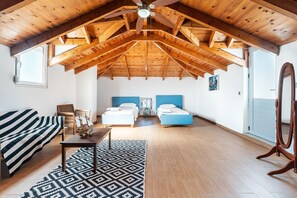 Un ático amplio con techo de madera inclinado, dos camas con cabeceros azules, una mesa de madera, una alfombra estampada y un ventanal grande.