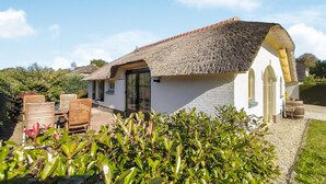Uma casa com telhado de palha, parede de tijolos e um pequeno jardim com arbustos verdes.