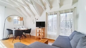 Uma sala de estar moderna com um sofá cinzento, uma mesa de jantar com cadeiras, uma TV sobre um suporte de madeira e uma janela ampla com persianas.