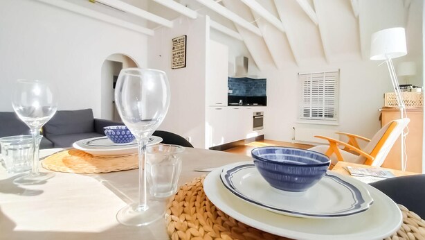 Uma área de refeições com uma mesa posta para dois, incluindo um jogo americano trançado, uma tigela azul e taças de vinho transparentes.