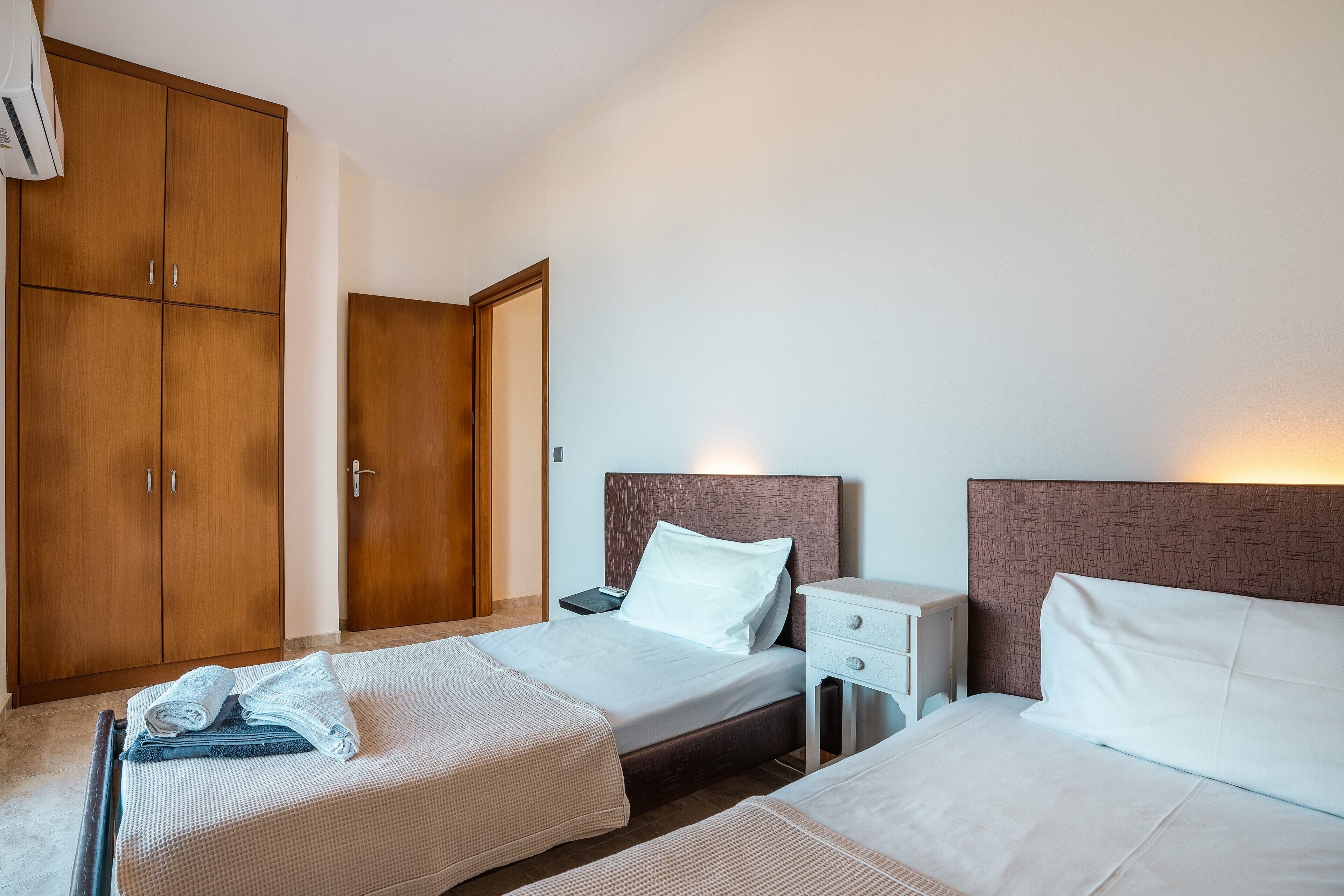A hotel room with two beds, wooden wardrobes, a bedside table with a lamp, and a wall-mounted air conditioner.