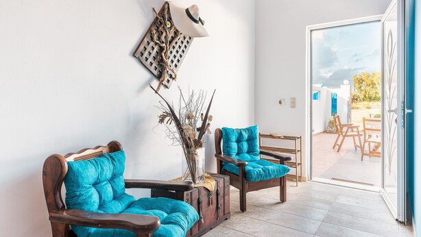 Une pièce comprenant deux chaises en bois avec des coussins turquoise, une commode en bois, un chapeau accroché au mur et une vue sur une terrasse extérieure, visible à travers une porte vitrée.