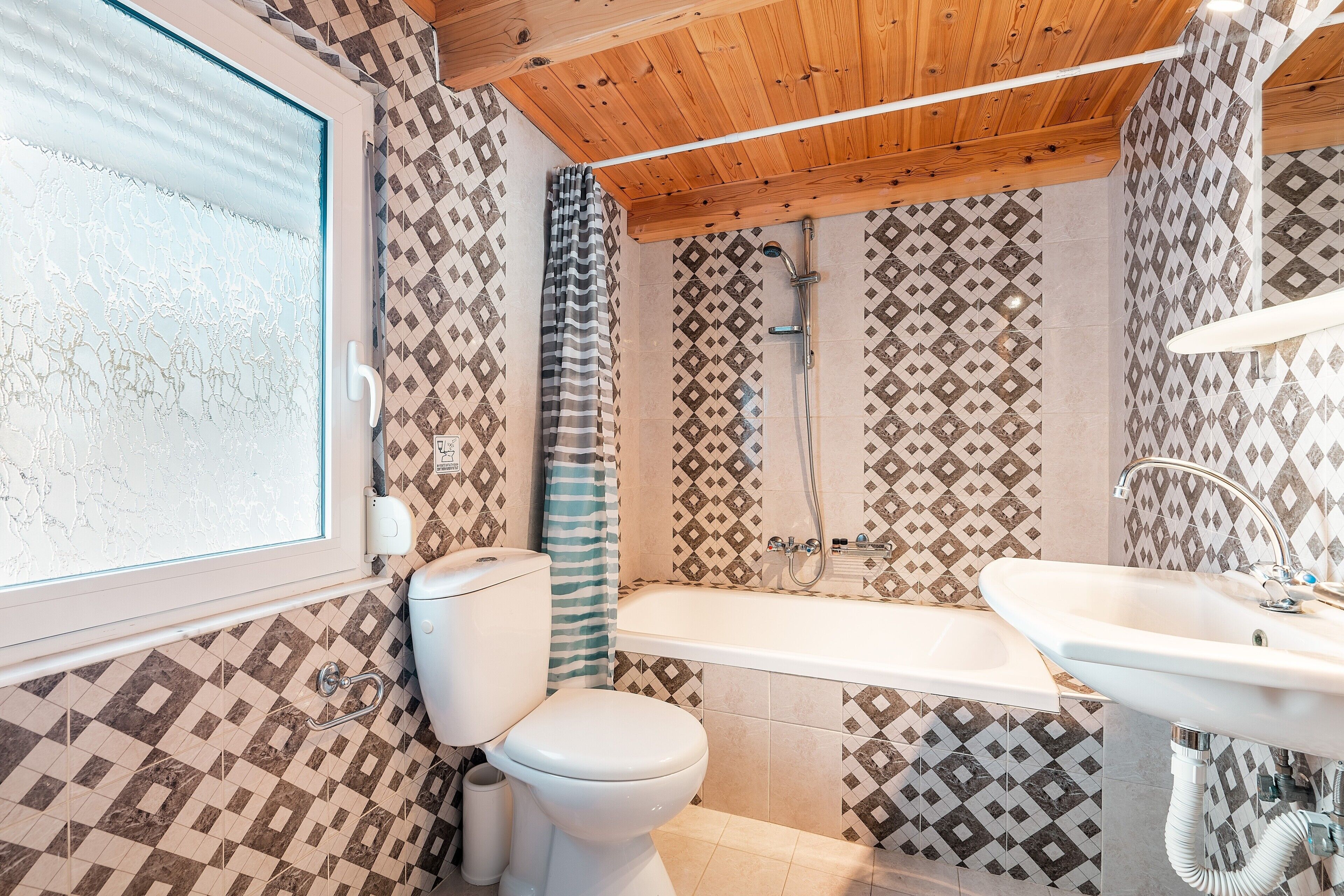 Un baño con inodoro, ducha y lavamanos, con baldosas estampadas y techo de madera.