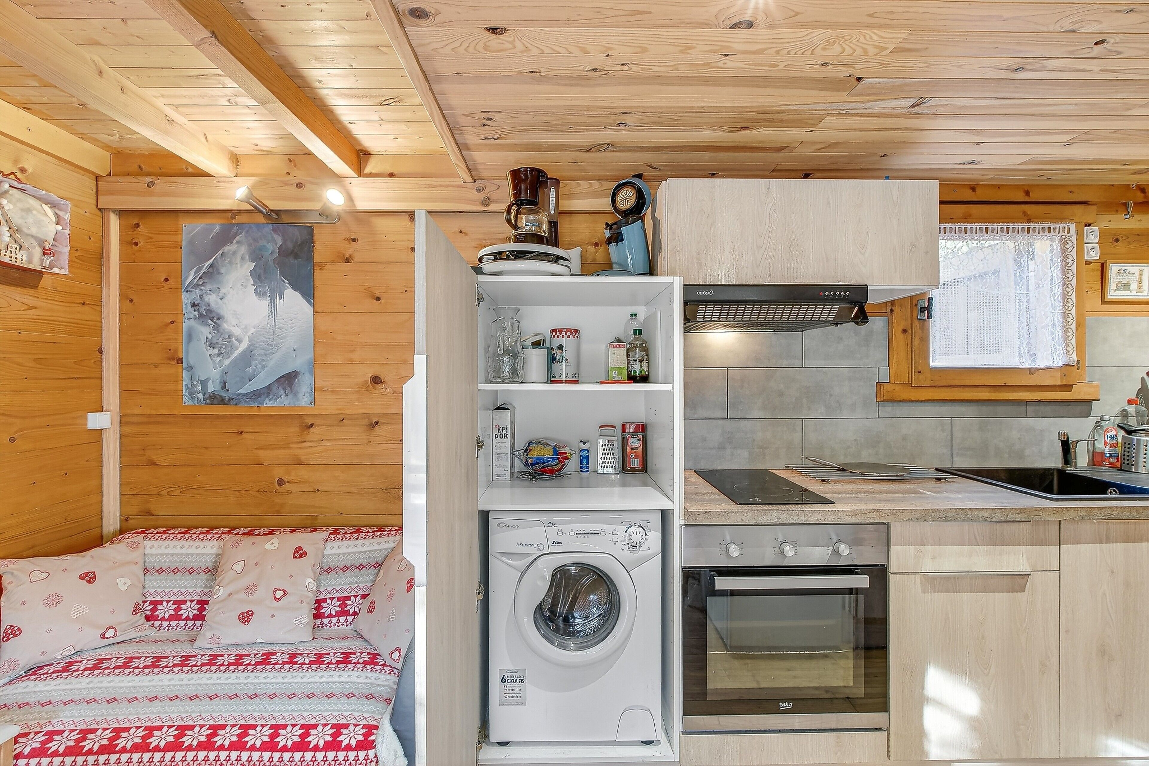 A compact room with a washing machine, a small kitchenette, a bed with patterned bedding, and a wooden ceiling.