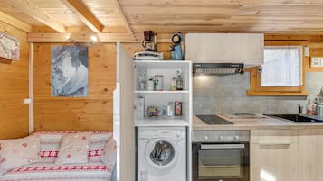 A compact room with a washing machine, a small kitchenette, a bed with patterned bedding, and a wooden ceiling.