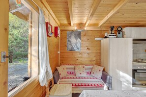 A wooden cabin interior with a bed, a small table, a window with curtains, a kitchen area, and a view of the outdoors.