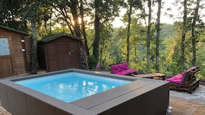 Un jacuzzi rectangulaire entouré d’une terrasse en bois, une cabane en bois et un fauteuil de relaxation avec un coussin rose.