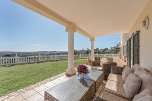 A covered outdoor area with a view of a white fence, a grassy lawn, and a distant landscape.
