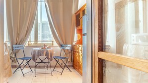 Une pièce avec une fenêtre, des rideaux, une table avec des chaises, un meuble et un réfrigérateur.