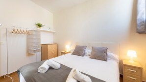 Un lit bien fait, recouvert d’une couverture grise, avec deux oreillers et une lampe de chevet. On y trouve une commode en bois sur laquelle est posée une plante, un porte-serviettes avec des serviettes pliées, et une petite étagère également ornée d’une plante.