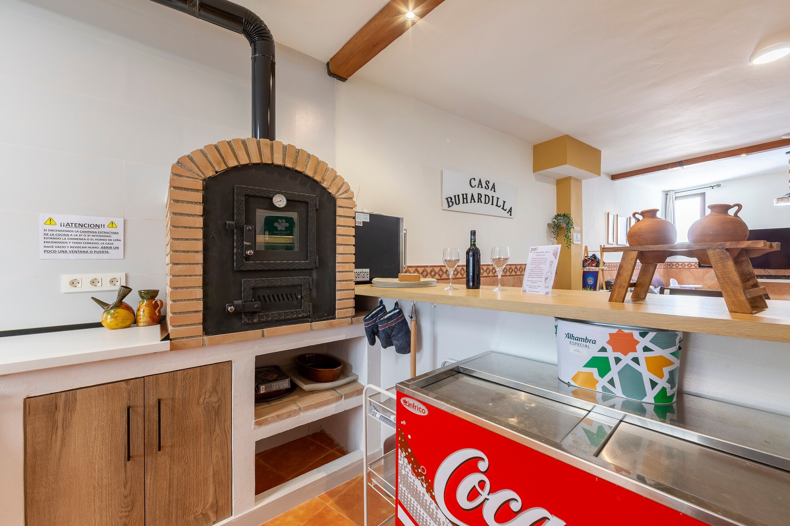 Une cuisine avec un four en briques, une enseigne indiquant « Casa Buhardilla », et un réfrigérateur à boissons de la marque Coca-Cola.