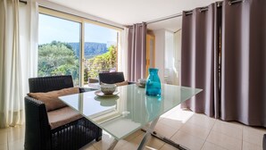 A room with a glass table, two wicker chairs, a blue vase, and a view of greenery and mountains.