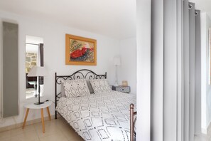 A bedroom with a bed, a round table with a lamp, a painting on the wall, and a window with curtains.