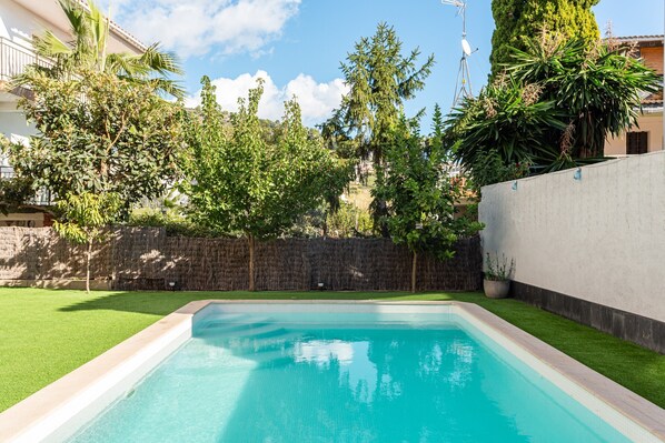 A rectangular swimming pool with a stone border, surrounded by a well-maintained lawn and a stone wall.