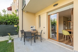 Um pátio com mesa e cadeiras, uma porta de vidro deslizante que dá para uma área de refeições interna, e uma cerca verde com plantas.