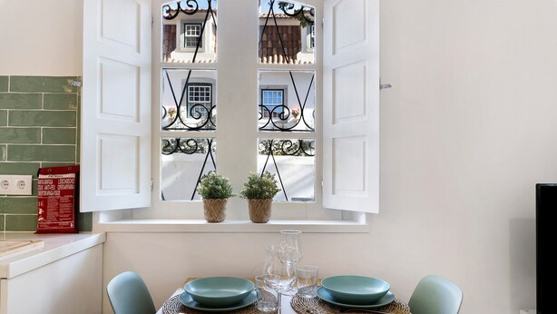 A dining area with a table set for four, a window with decorative iron bars, and a view of a building outside. - Apartment 'Jardim Da Parada' with Wi-Fi and Air Conditioning (Cascais)