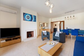 A modern living room with a flat-screen TV, a fireplace, a dining area with blue chairs, and a large abstract painting on the wall. - Apartment 'Duo Village' with Shared Pool, Wi-Fi and Air Conditioning (Alvor)