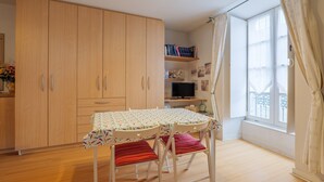 Ein Zimmer mit Holzschrank, Tisch mit Stühlen, Fernseher und einem Vorhangfenster.