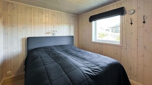 A bedroom with a dark-colored bed, wooden walls, a window with a black valance, and a small wall-mounted light fixture.